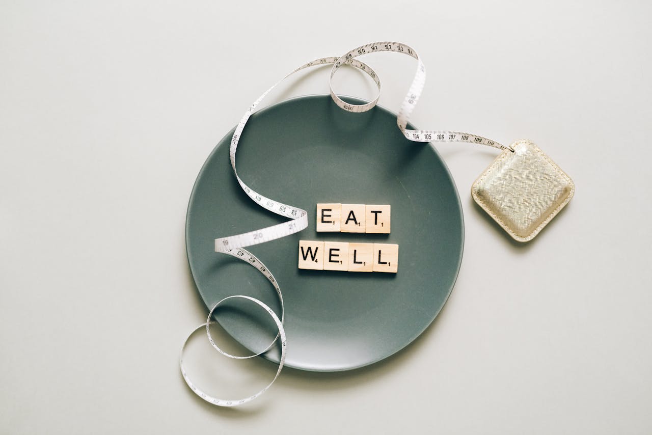 Flat lay of scrabble tiles spelling 'eat well' on a plate with a tape measure, promoting healthy eating.